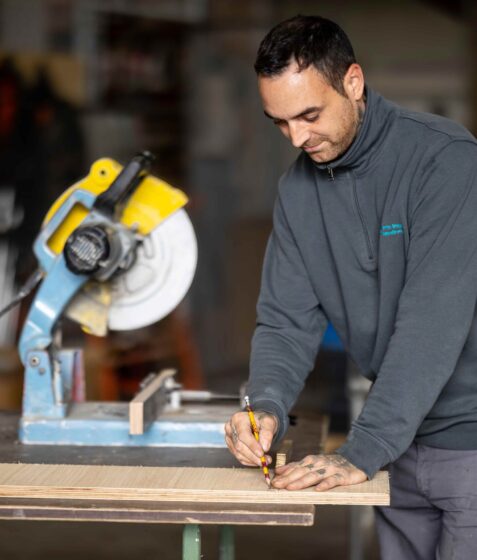 Handwerker in einer Werkstatt markiert ein Holzbrett mit einem Bleistift neben einer Kappsäge.